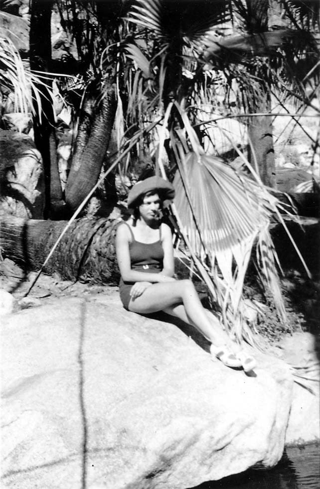 A portrait of Elisabeth "Betty" Hall wearing a wide-brimmed hat and swimsuit, sitting on a large rock surrounded by palm trees and rugged terrain.
