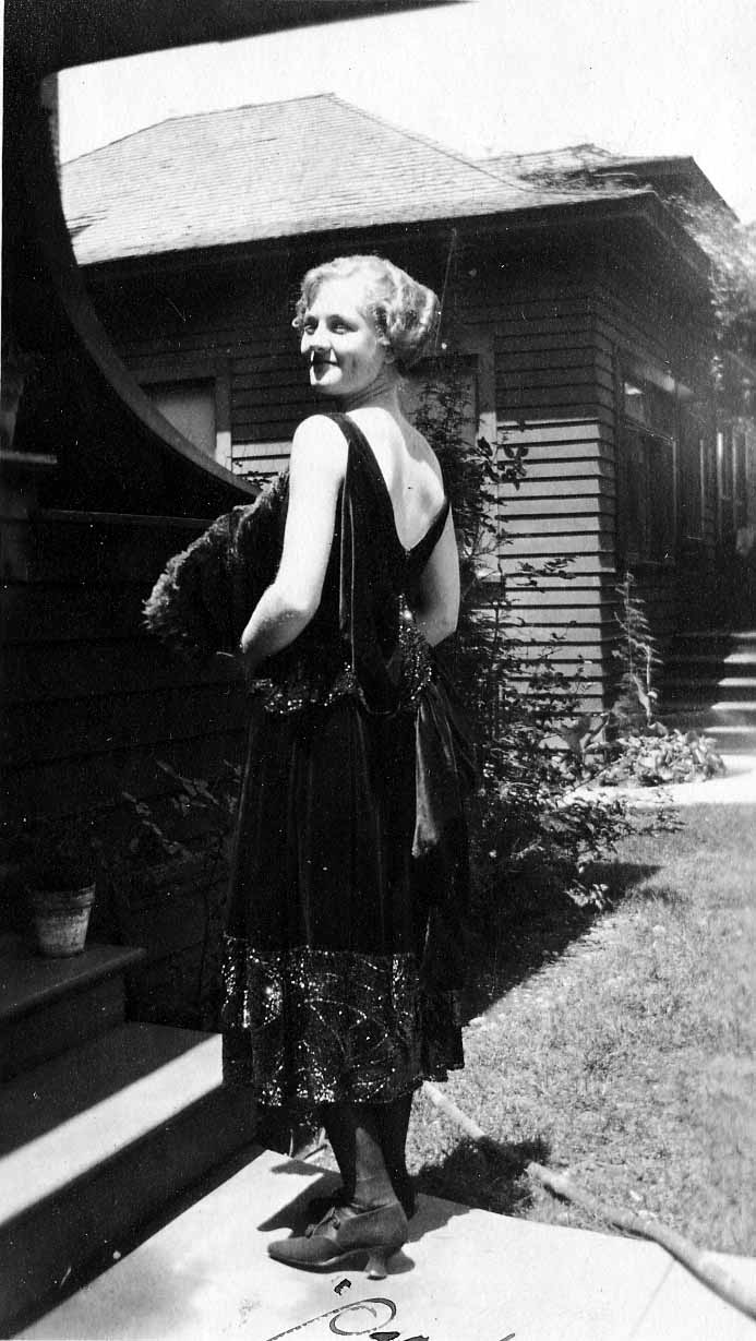 A glamorous woman in a backless beaded 1920s evening dress and heels holding a large feather fan on porch steps during a wedding celebration.