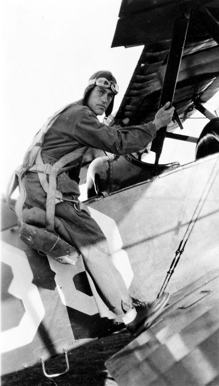 An action shot of a flight cadet wearing a parachute harness, stepping up onto the wing to climb into the cockpit of a training biplane.