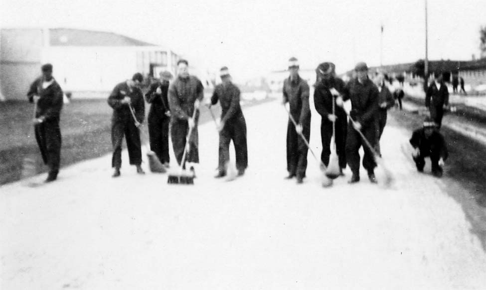A group of Army Air Corps flight cadets in uniform sweeping a wide dirt or gravel road with push brooms.