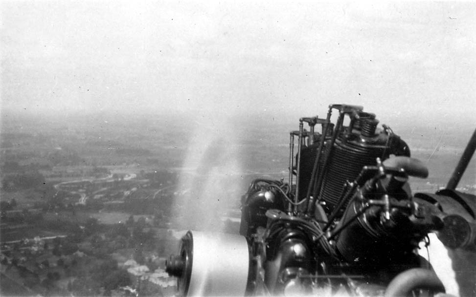 An over-the-shoulder air-to-air view looking past the spinning propeller and exposed cylinders of an aircraft engine down toward the landscape below.