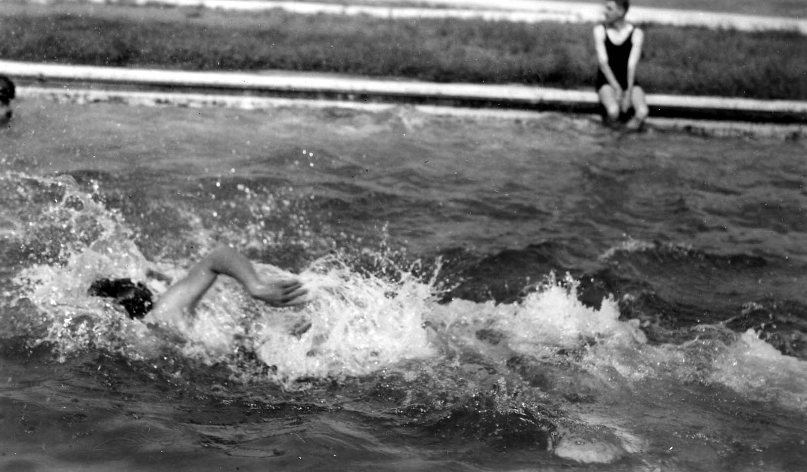 A person swimming freestyle in a pool with water splashing, while another person in a dark swimsuit sits on the edge in the background.