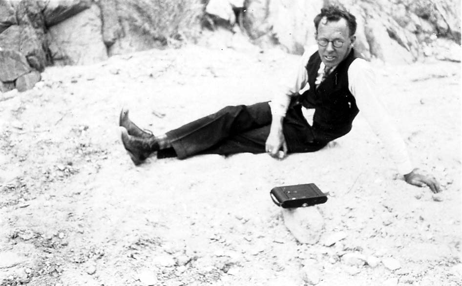 A man wearing round spectacles, a vest, and tie lounging casually on a rocky dry slope with a vintage folding roll-film camera resting on a stone beside him.