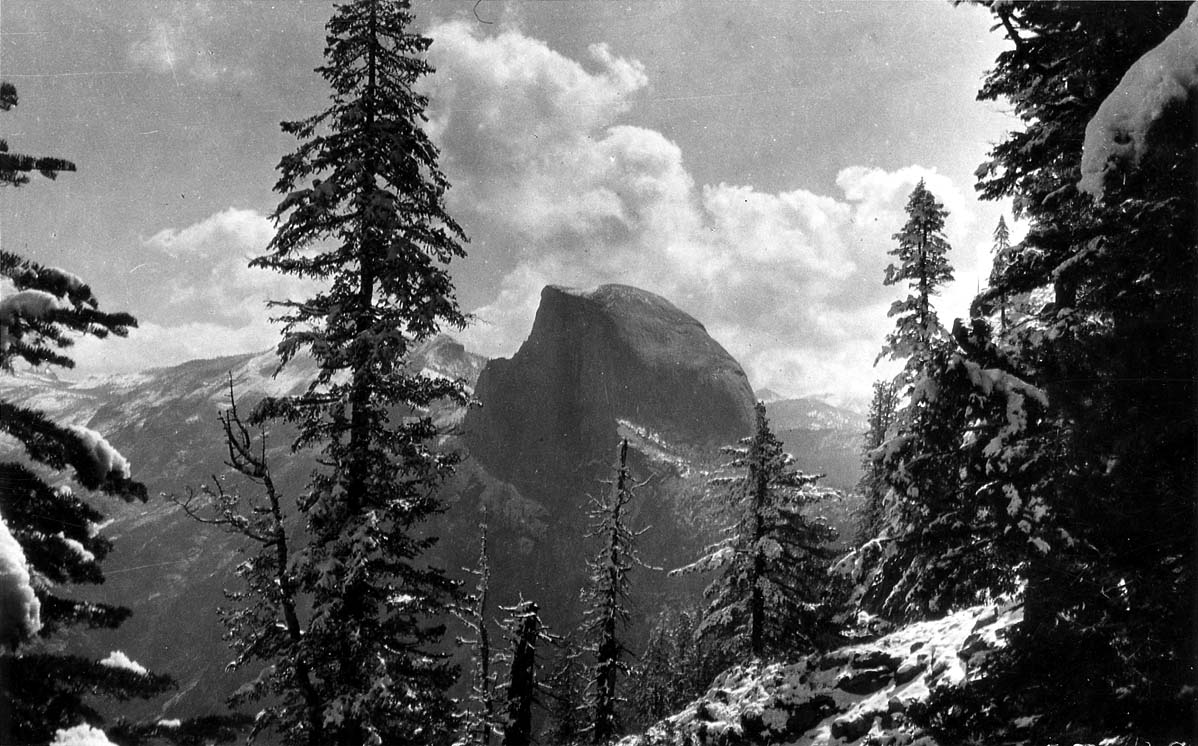 The sheer granite monolith of Half Dome rising in the distance, framed by snow-covered conifers under billowing cumulus clouds.