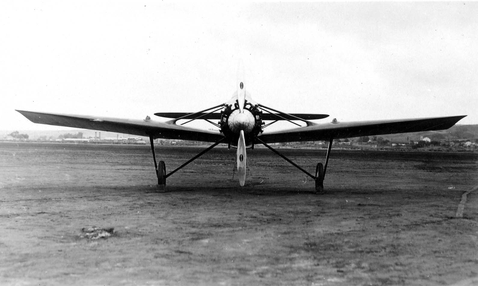 A symmetrical head-on frontal view of the Hall X-1 tandem-wing prototype on a wide dirt airfield, showing its engine, propeller, and struts.