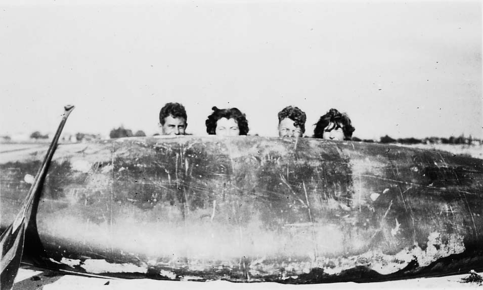Four people humorously peering over the top of an overturned dark canoe on a sandy beach.