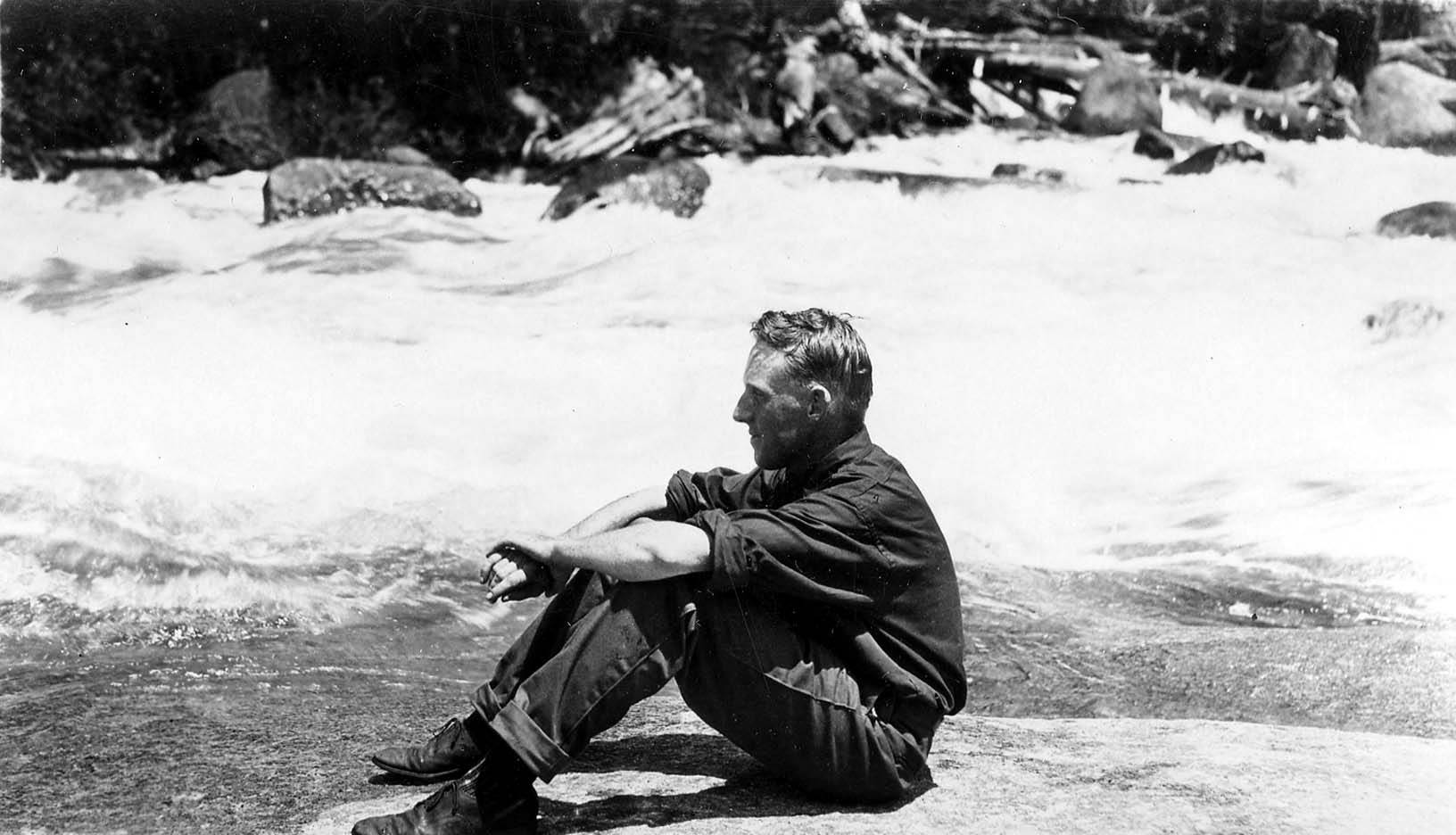 A man in work clothes seated profile on a flat granite boulder beside a churning mountain river with foaming white water rapids.