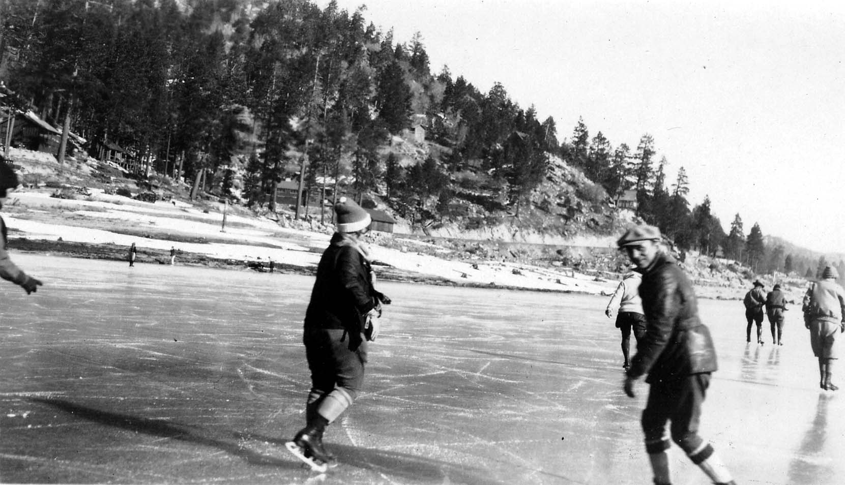A large group of men and women wearing winter clothing and ice skates, standing together near the snowy bank of a frozen lake.