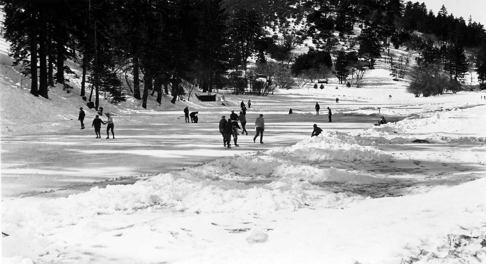 A wide view of a frozen lake dotted with ice skaters and snow banks, with long, stark shadows cast by the afternoon sun.