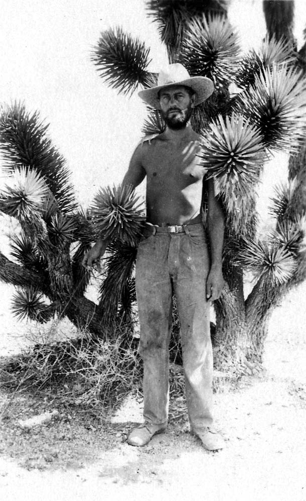 A shirtless Donald A. Hall Sr. wearing a straw hat and jeans, standing in front of a large Joshua tree.