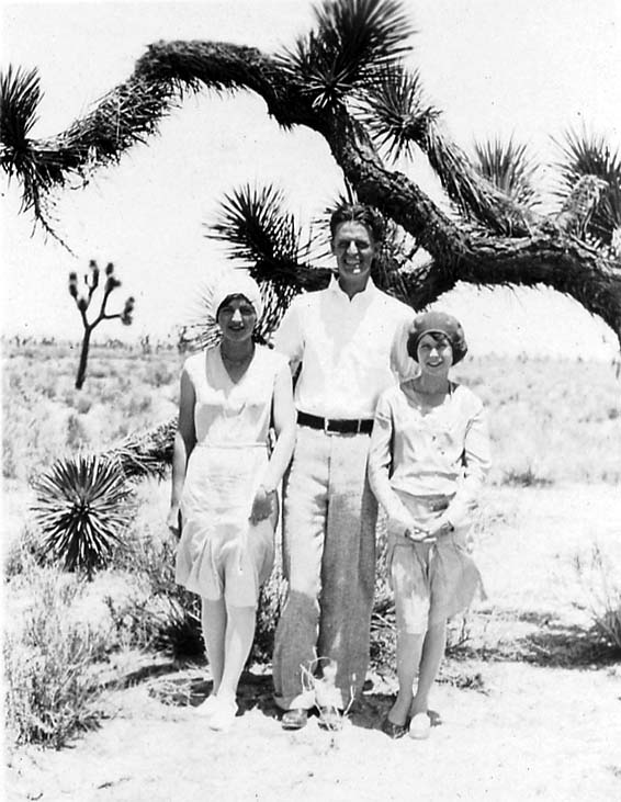A man and two women in light 1920s summer attire posing together under the twisting limbs of a large Joshua tree in the arid Mojave Desert.