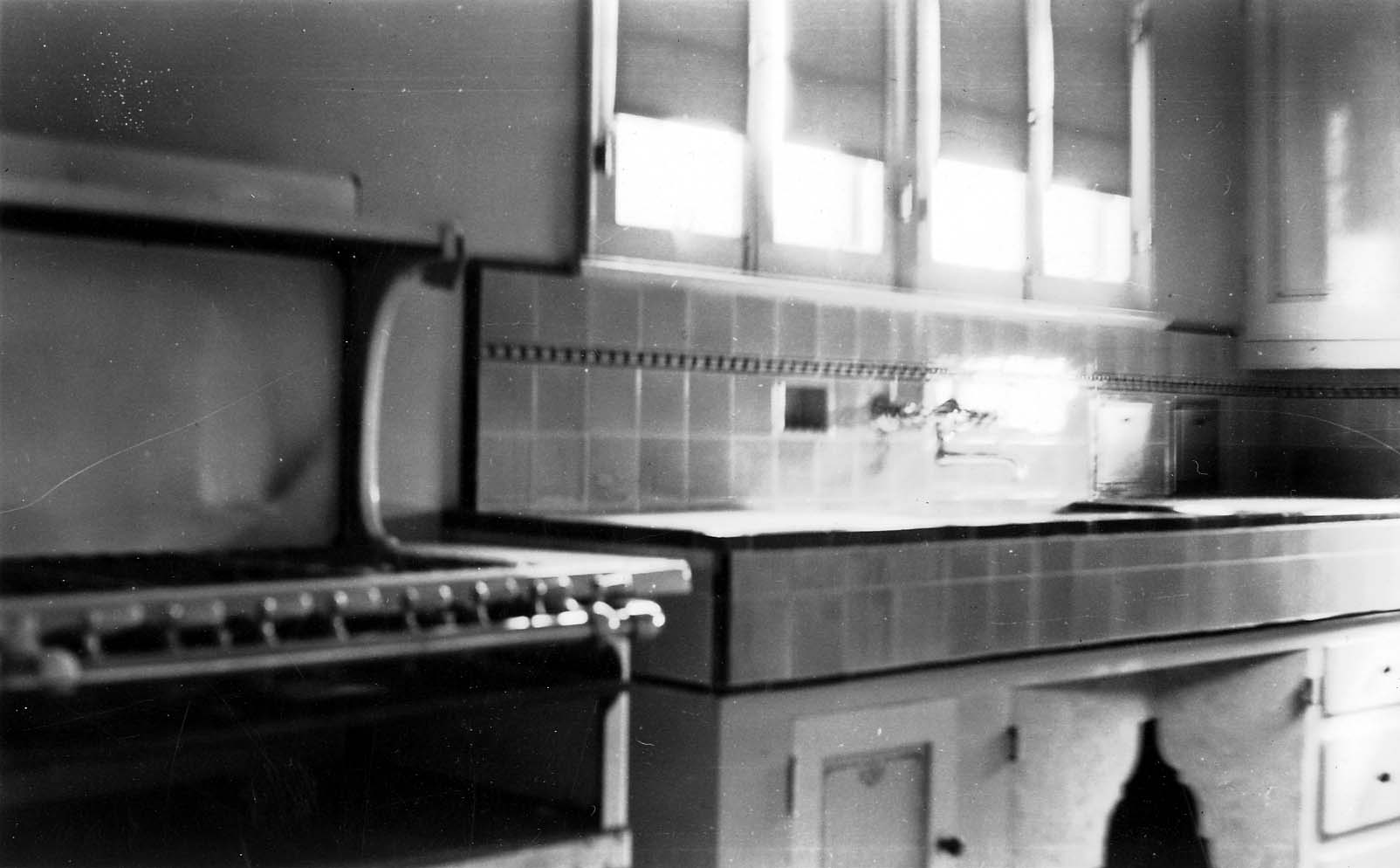 An interior view of a vintage 1920s kitchen, highlighting a large gas stove, tiled countertops, and sunlight streaming through the windows.