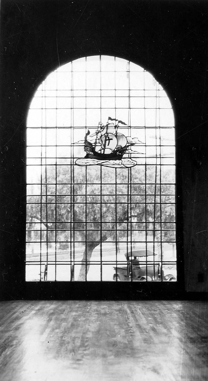 An interior view looking out through a massive, arched window featuring a stained-glass ship motif, with a vintage car visible parked outside.
