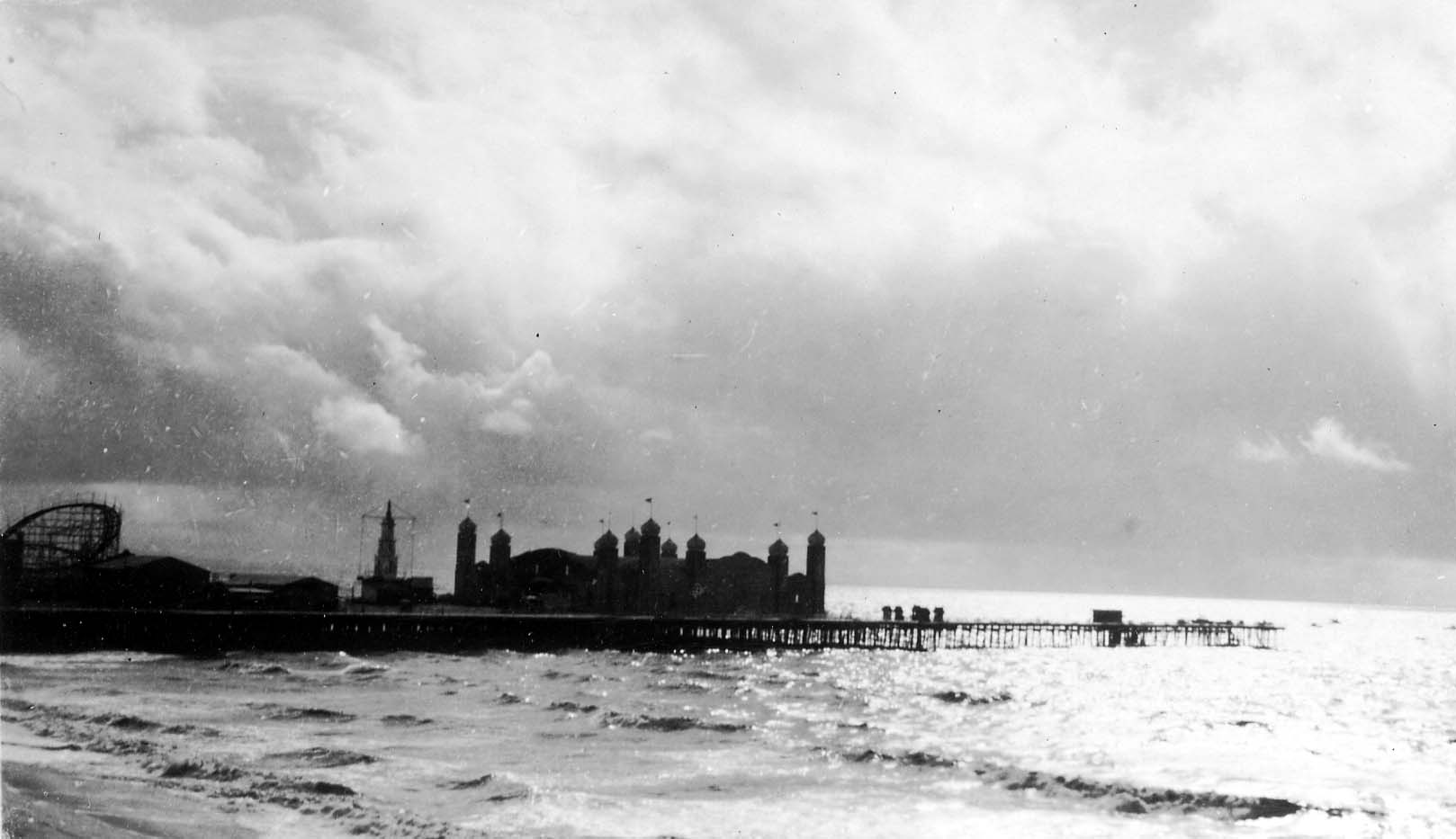 A wide, atmospheric view of the massive La Monica Ballroom building sitting at the end of the Santa Monica Pier over choppy ocean waves beneath a cloudy sky.