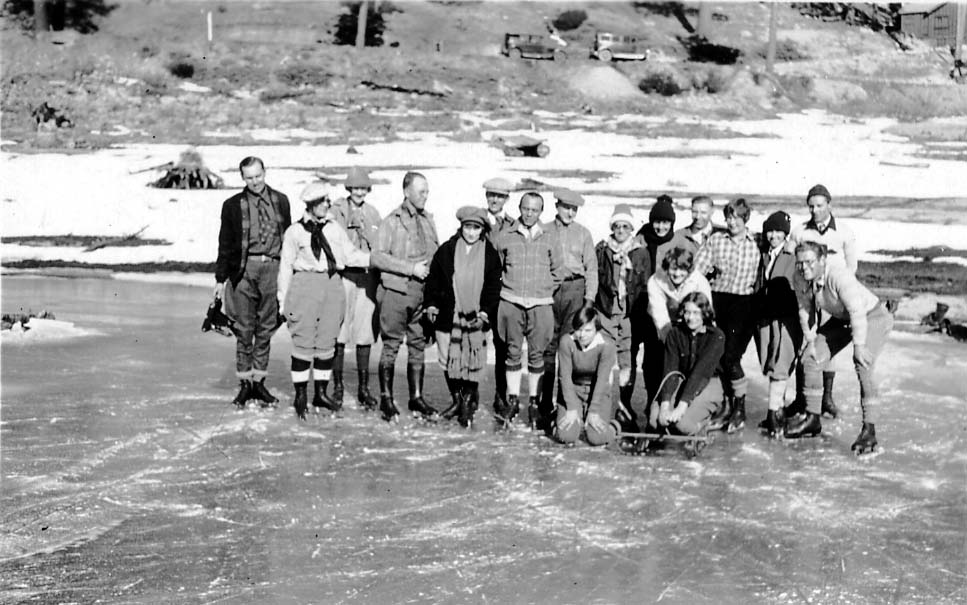 A large, mixed group of men and women wearing winter gear and ice skates, posing together for a group portrait on a frozen lake.