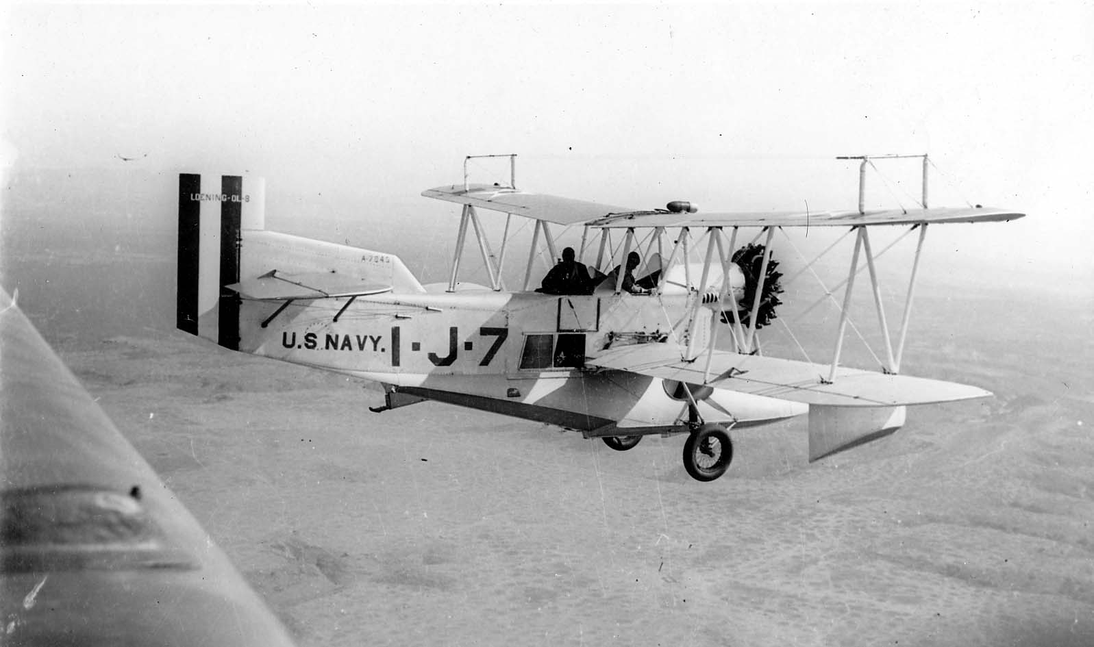 An air-to-air side profile of a U.S. Navy Loening OL-8 amphibian biplane serial A-7845 flying over the San Diego landscape.
