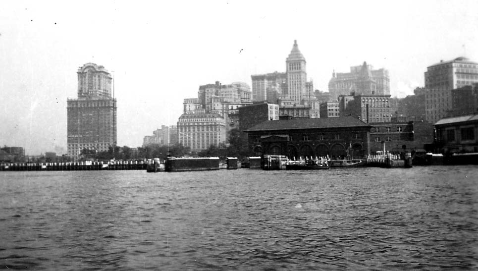 A harbor view of the Lower Manhattan skyline from the water, featuring the arched Pier A fireboat station, the Whitehall Building, and the Standard Oil Building.