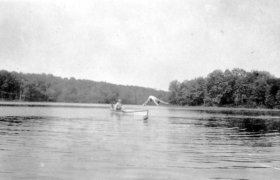 A man diving headfirst into a lake from the edge of a small wooden rowboat, while another man sits in the boat watching.