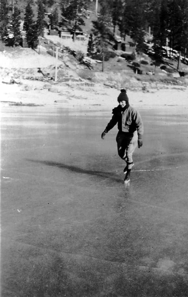A man wearing a knit cap and a heavy jacket ice skating gracefully across a frozen lake, with a snowy, wooded hillside in the background.