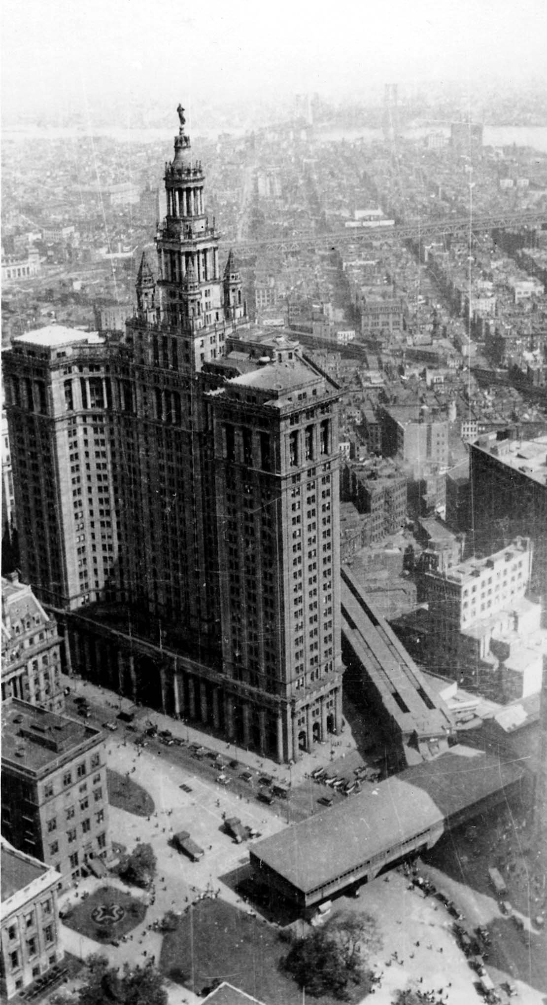 An elevated perspective looking directly at the colonnaded tower and gilded Civic Fame statue of the Manhattan Municipal Building above Park Row terminal.