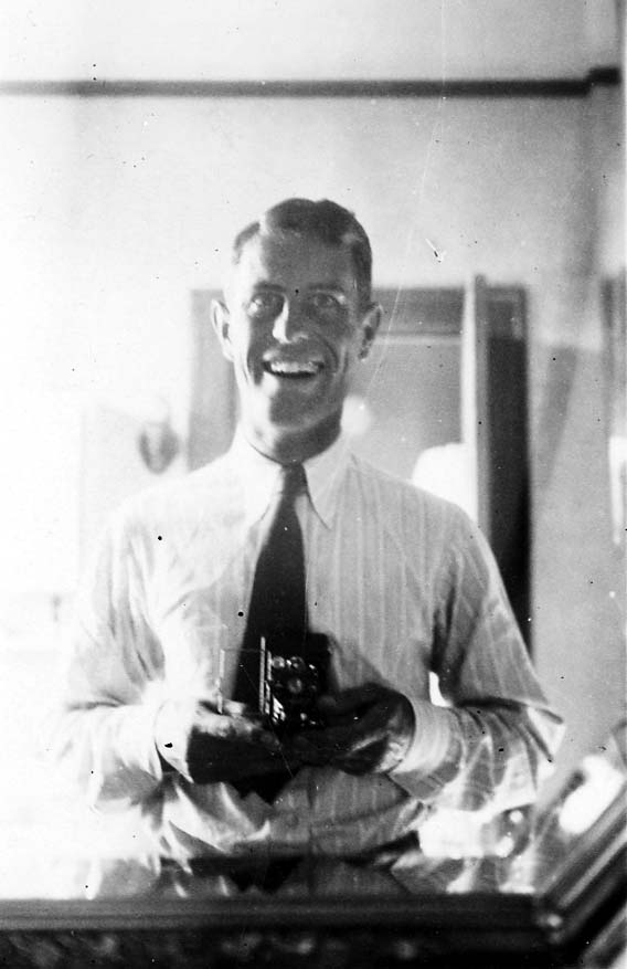 A smiling Donald A. Hall Sr. taking a photograph of himself in a mirror using a vintage folding box camera, wearing a shirt and tie.