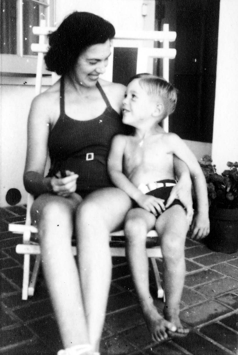 A woman and a young boy, both wearing swimsuits, sitting together in a white chair on a tiled patio and smiling at each other.