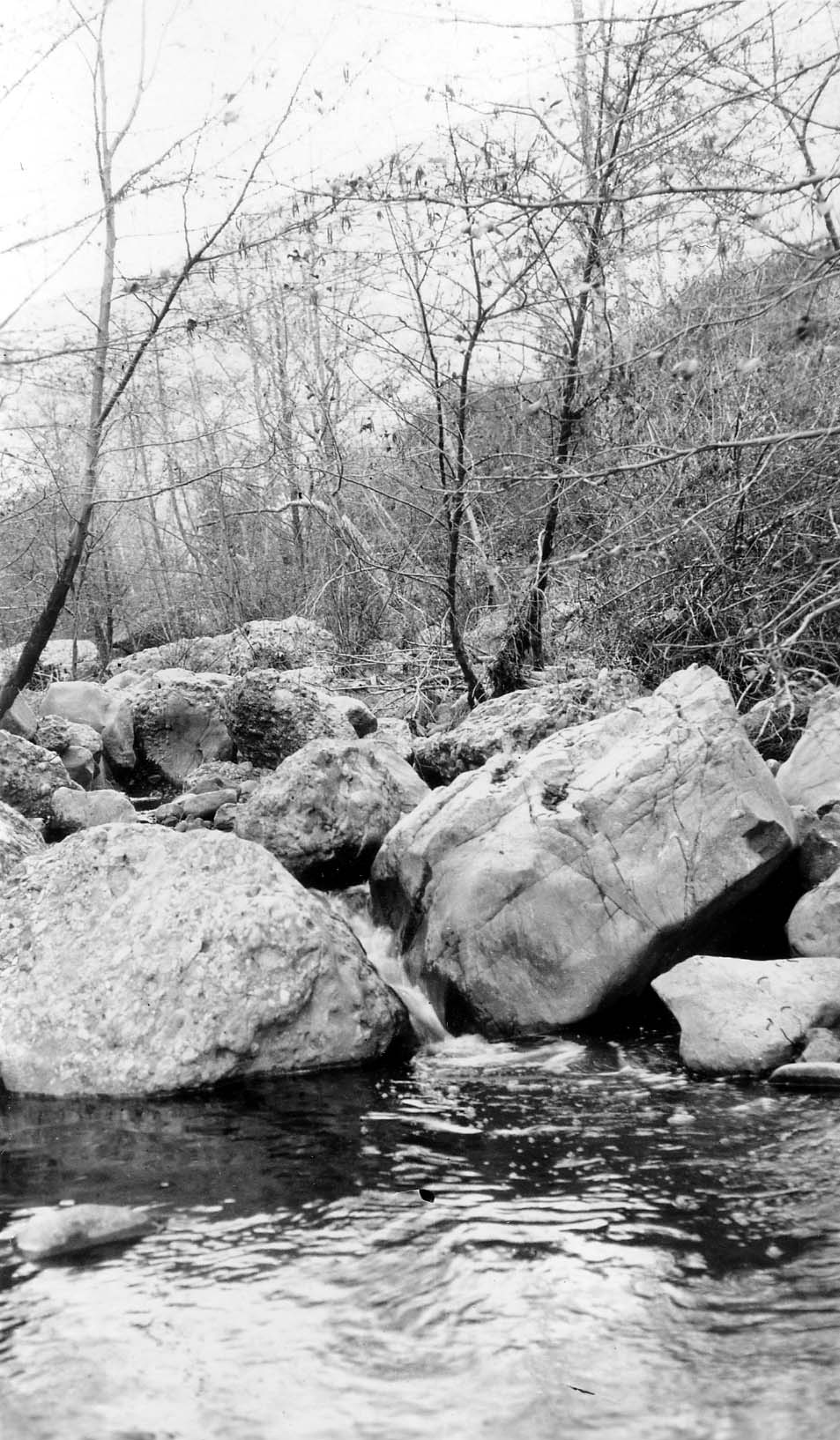A clear mountain stream flowing between large, weathered granite boulders and bare deciduous trees along the riverbank.