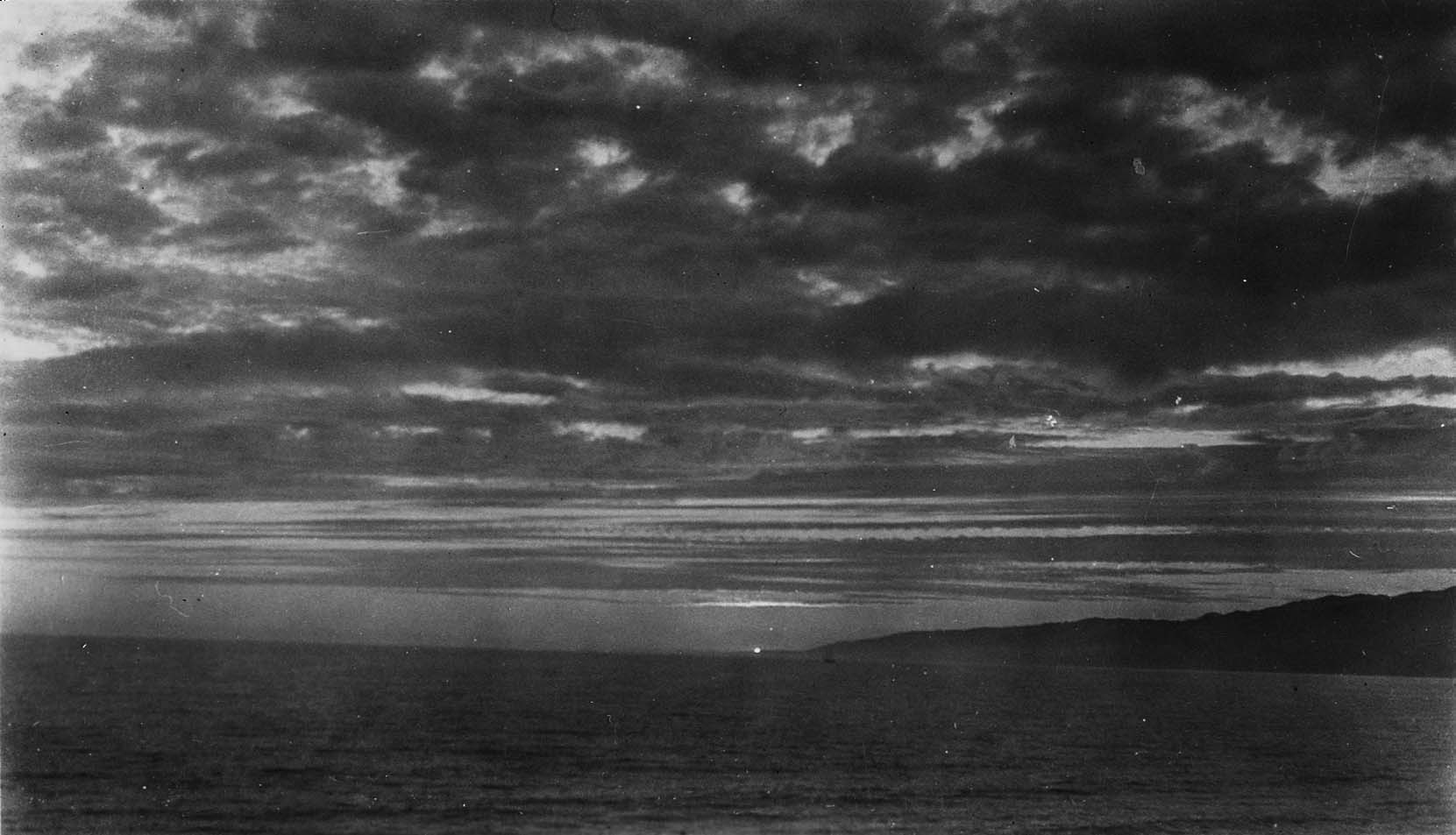 The setting sun glowing faintly on a flat Pacific Ocean horizon beneath dense, dark clouds with the silhouette of a coastal headland on the right.