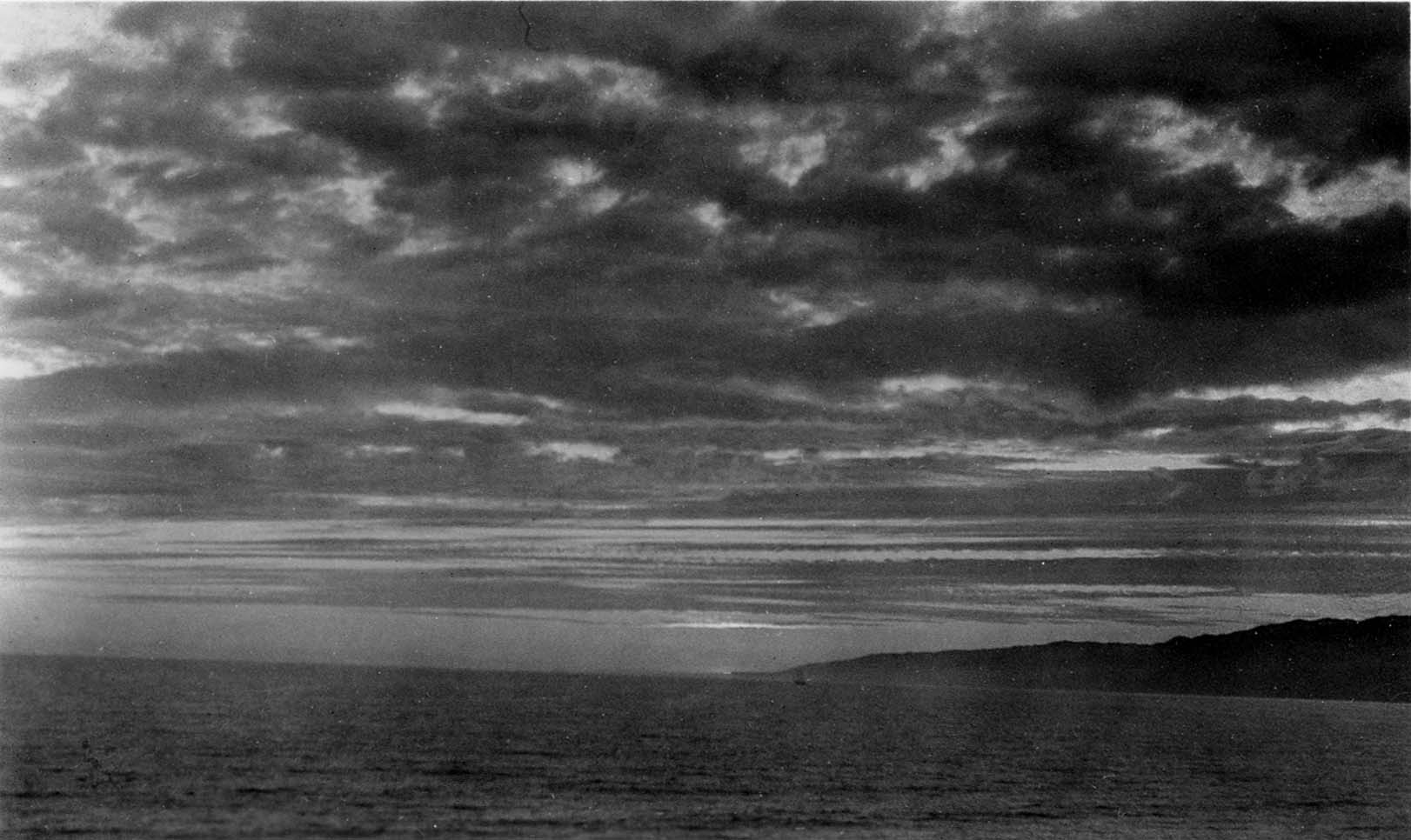 A dramatic Pacific Ocean sunset with layered, dark storm clouds stretching over calm waters toward a distant coastal headland.