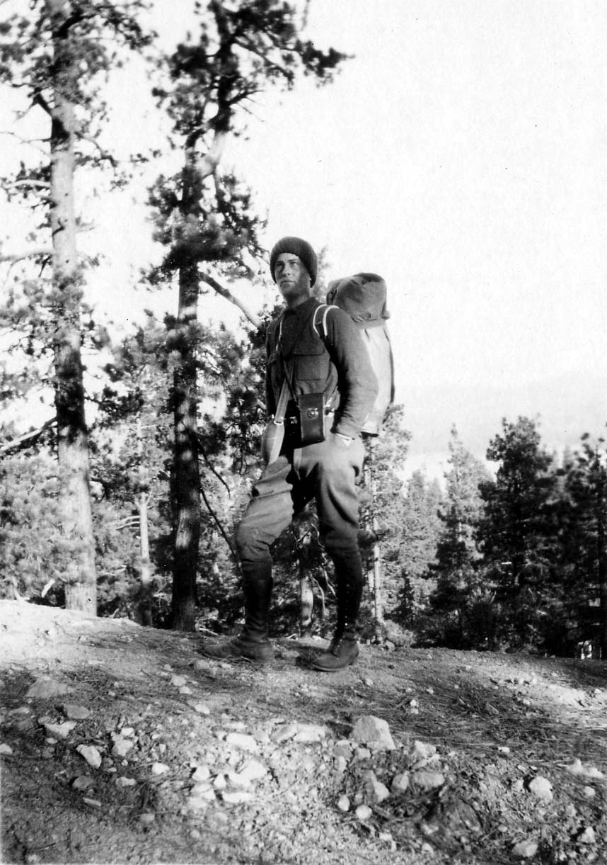 Donald A. Hall Sr. standing on a pine-covered ridge wearing a heavy canvas backpack, tall boots, and a knit winter cap.