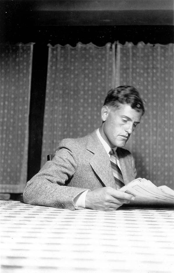 Donald A. Hall Sr. sitting indoors at a table, reading a newspaper while wearing a dapper suit and striped tie.