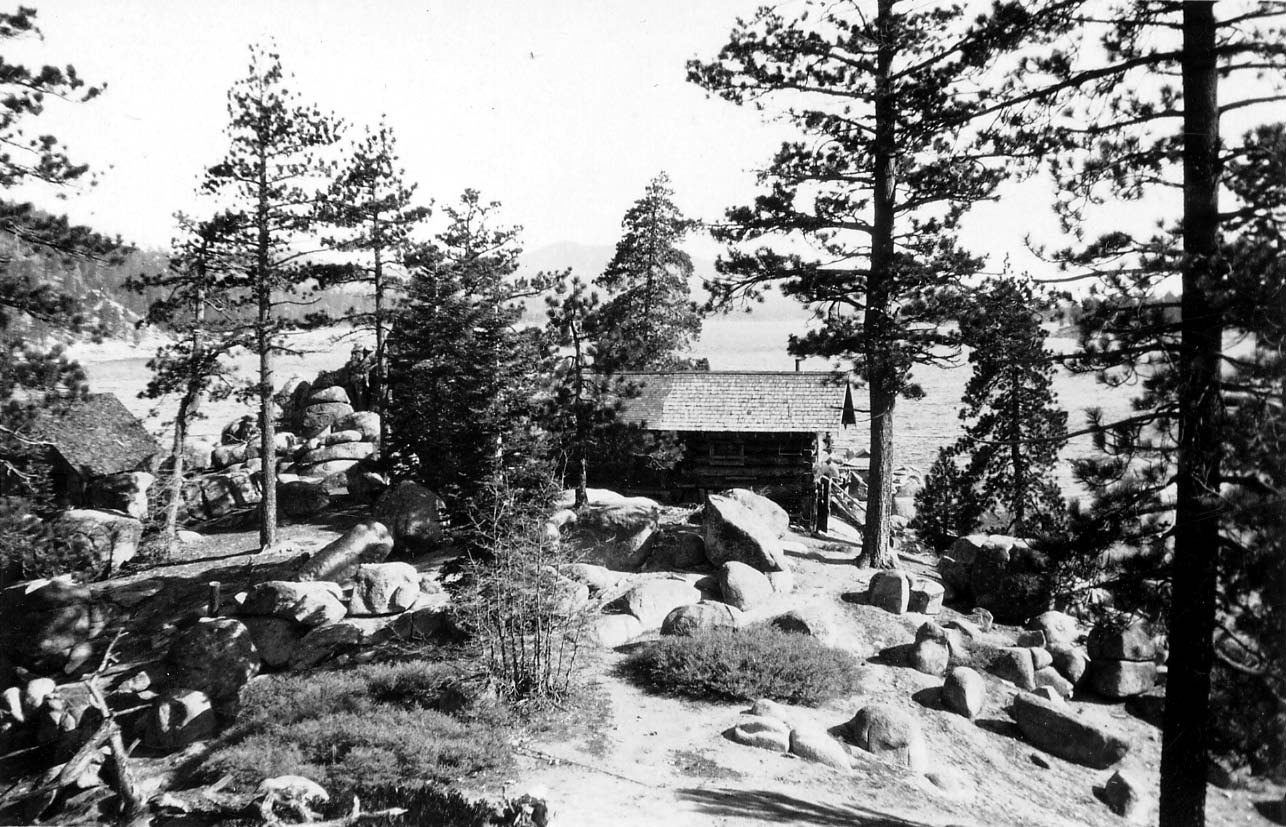 A rustic log cabin nestled among massive granitic boulders and tall pine trees on the rugged shore of a mountain lake.