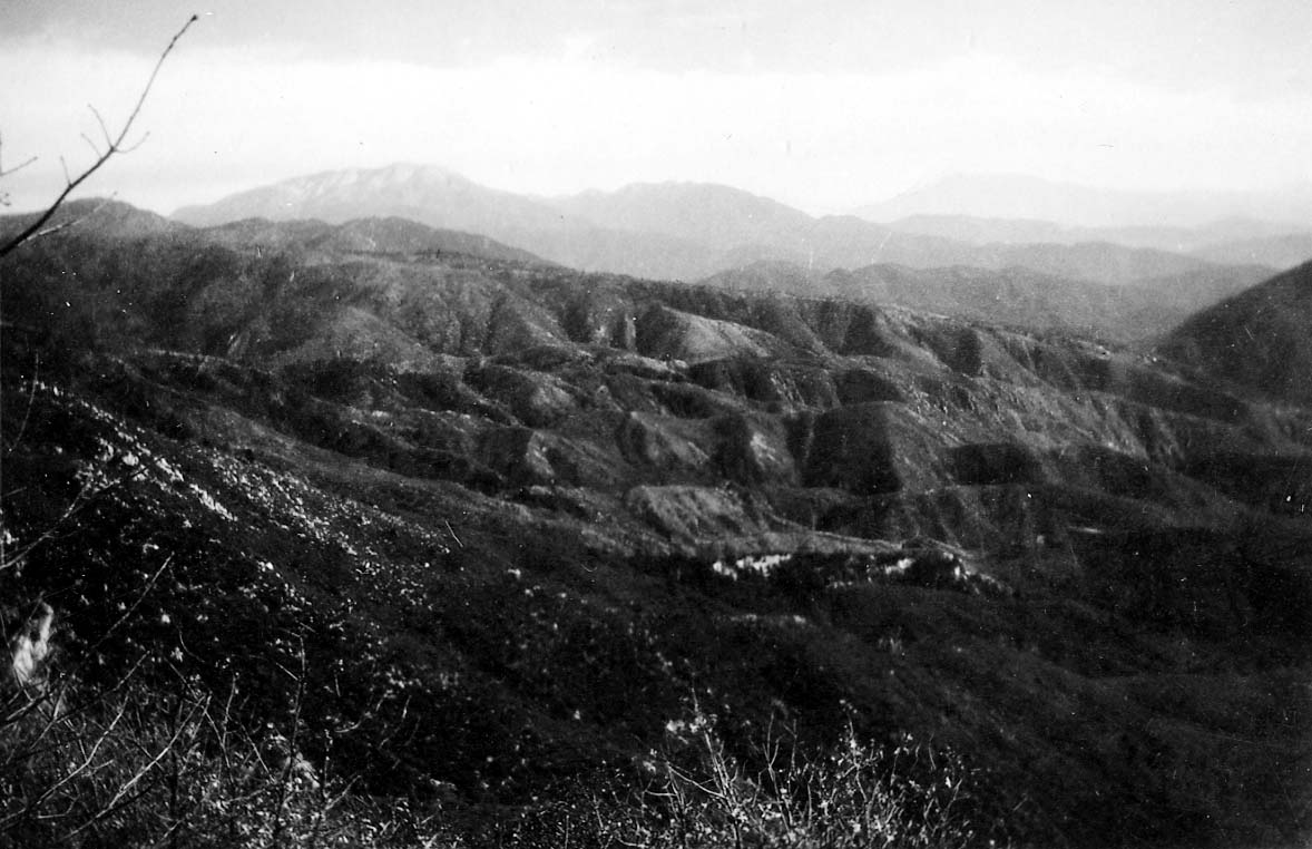 A high-altitude panoramic view of rolling, heavily creased mountain ridges receding into distant hazy peaks under an overcast sky.