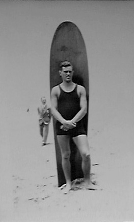 Donald A. Hall in tank swimsuit standing with a 10-foot solid wood surfboard on a San Diego beach.