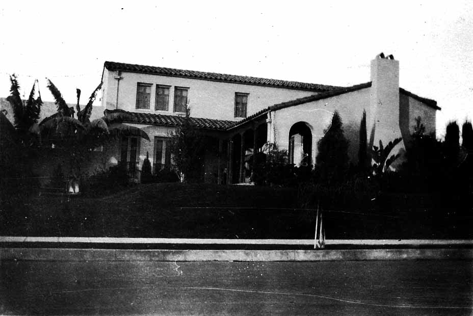 An elegant two-story Spanish Colonial Revival estate featuring white stucco walls, red tile roofs, an arched entryway, and tropical landscaping of palms and cypress trees.