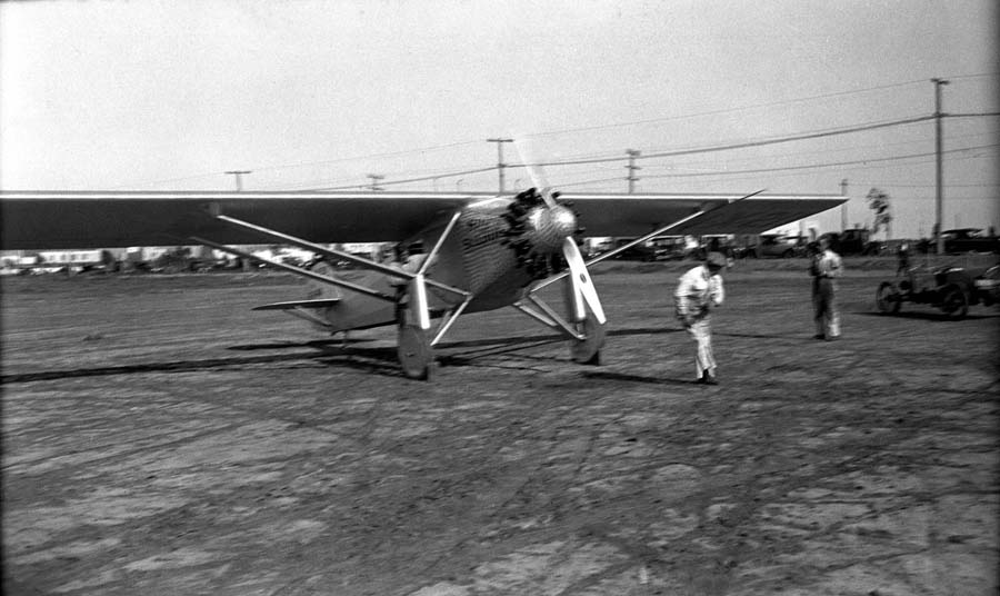 A mechanic pulling the propeller to start the engine of the Spirit of St. Louis on the dirt runway at Dutch Flats, with spectators watching.
