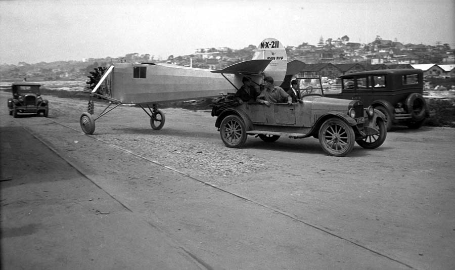 The Spirit of St. Louis being towed backward by a vintage car down a dirt road toward Dutch Flats, with a man looking back at the fuselage.