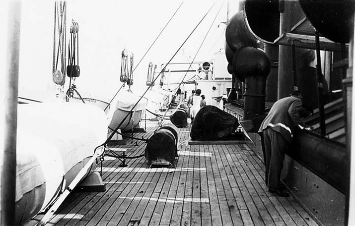 The wooden deck of a large steamship with canvas-covered lifeboats, large black ventilator cowls, and passengers looking over the railing.