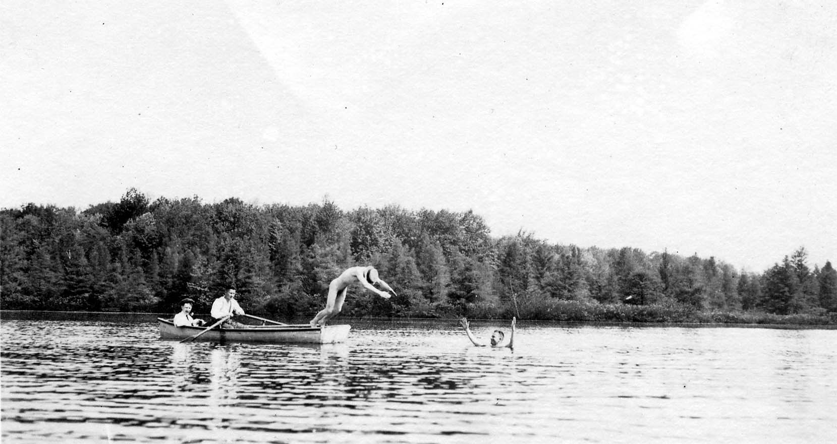 A swimmer diving into a lake from a rowboat, while another swimmer waves from the water and a third man watches from the boat.