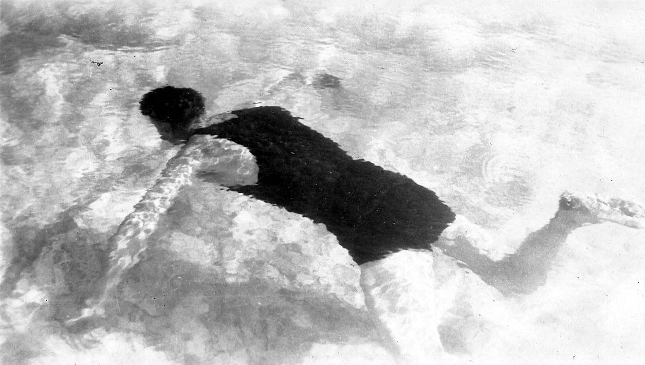 A high-angle photograph looking down through the surface of the water at a man gliding just beneath the surface.