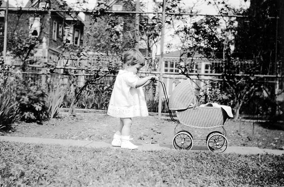 A young toddler in a white dress pushing a vintage woven wicker baby carriage along a garden walkway.