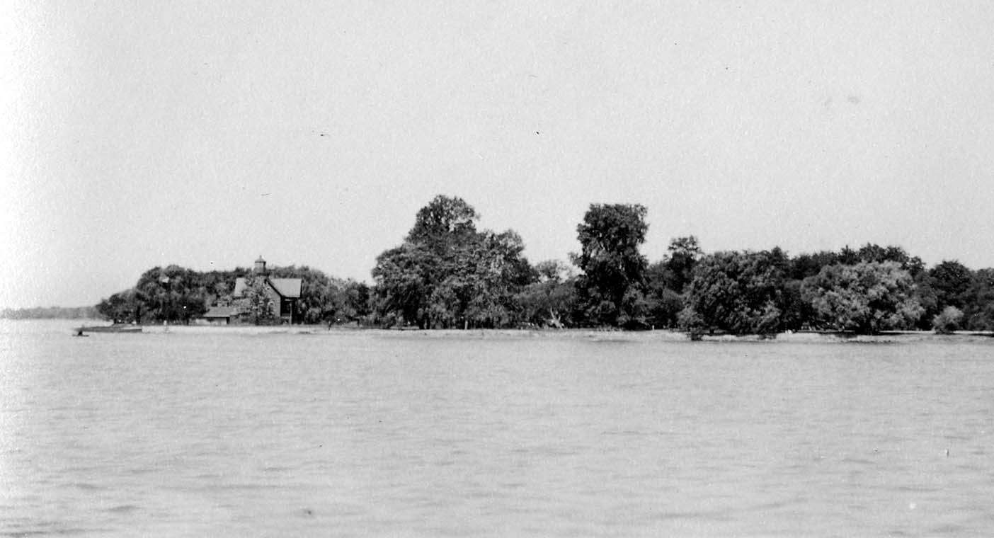 A wide view across calm freshwater toward a densely wooded island sheltering a small cottage beneath mature deciduous trees.