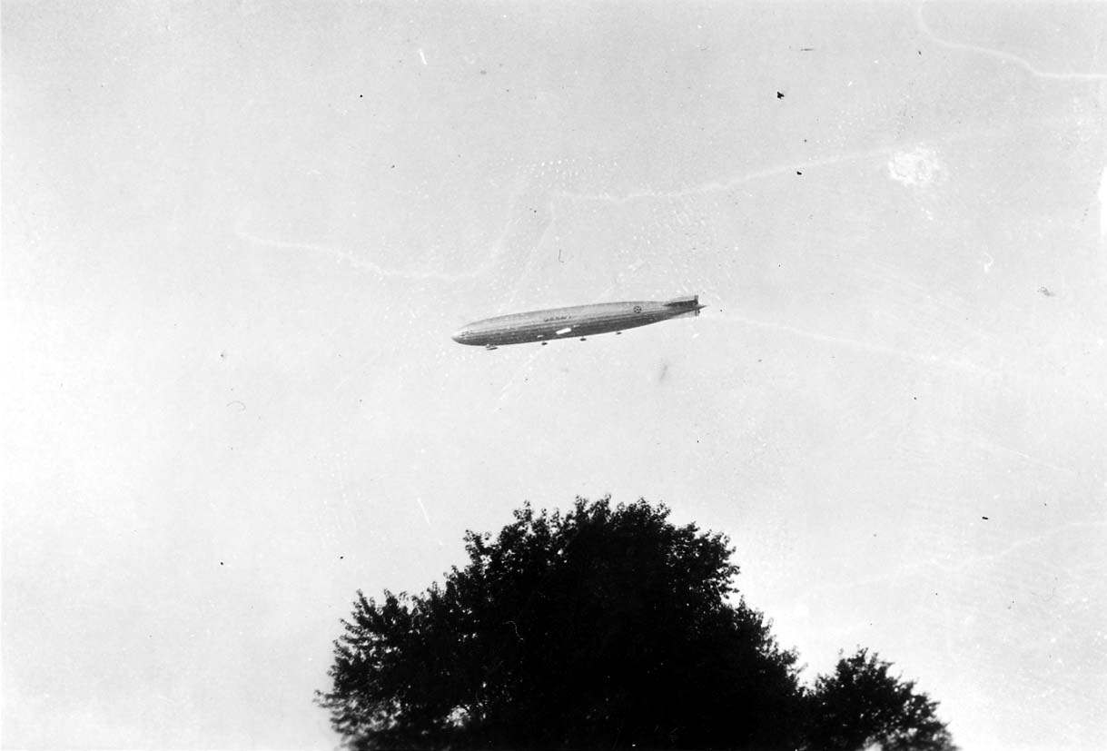 A distant aerial view of the U.S. Army non-rigid dirigible TC-2 cruising high above a large deciduous tree silhouette.