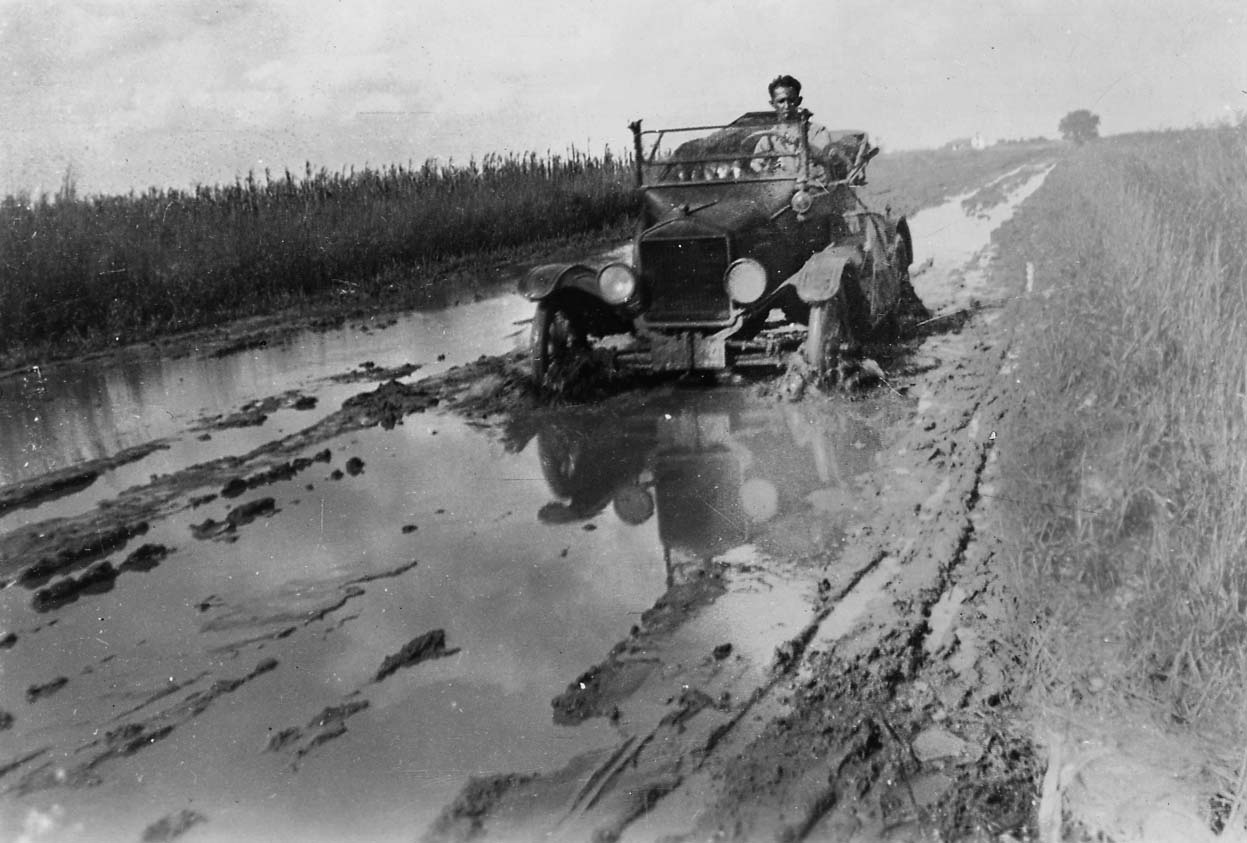 A vintage open-cab automobile, likely a Model T, buried to its axles in a deep, water-filled mudhole on a rural dirt road.