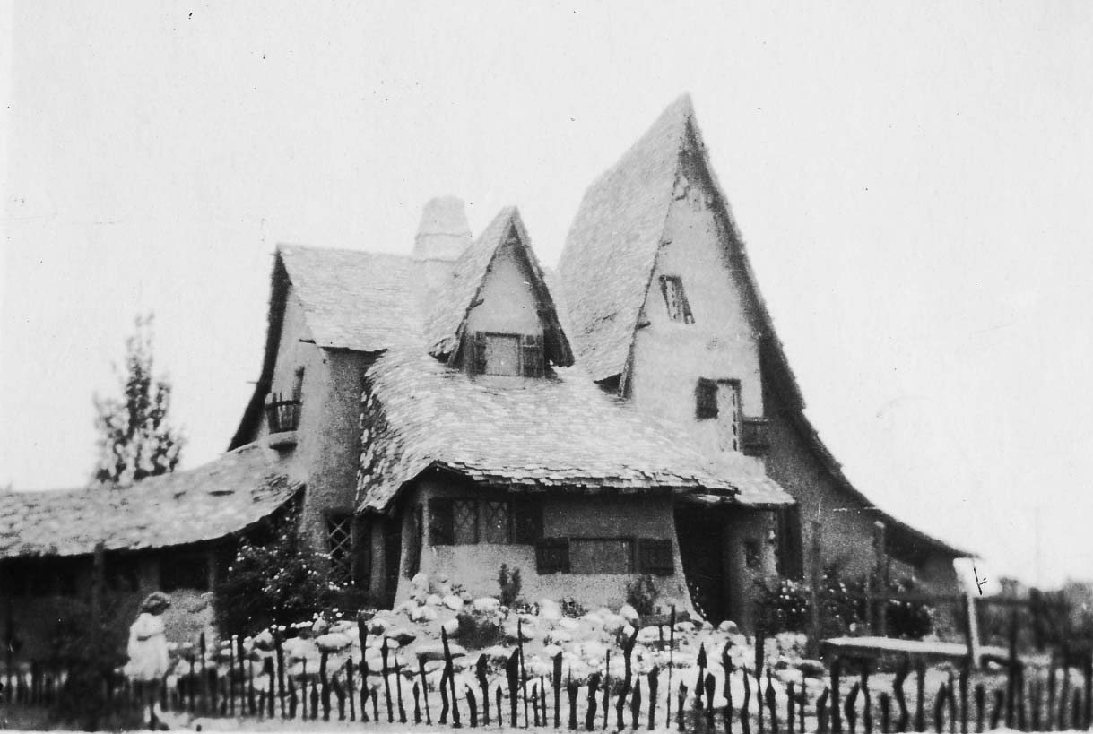 A whimsical, storybook cottage featuring dramatic, sharply peaked roofs and a rustic fence, with a small child standing in the front yard.