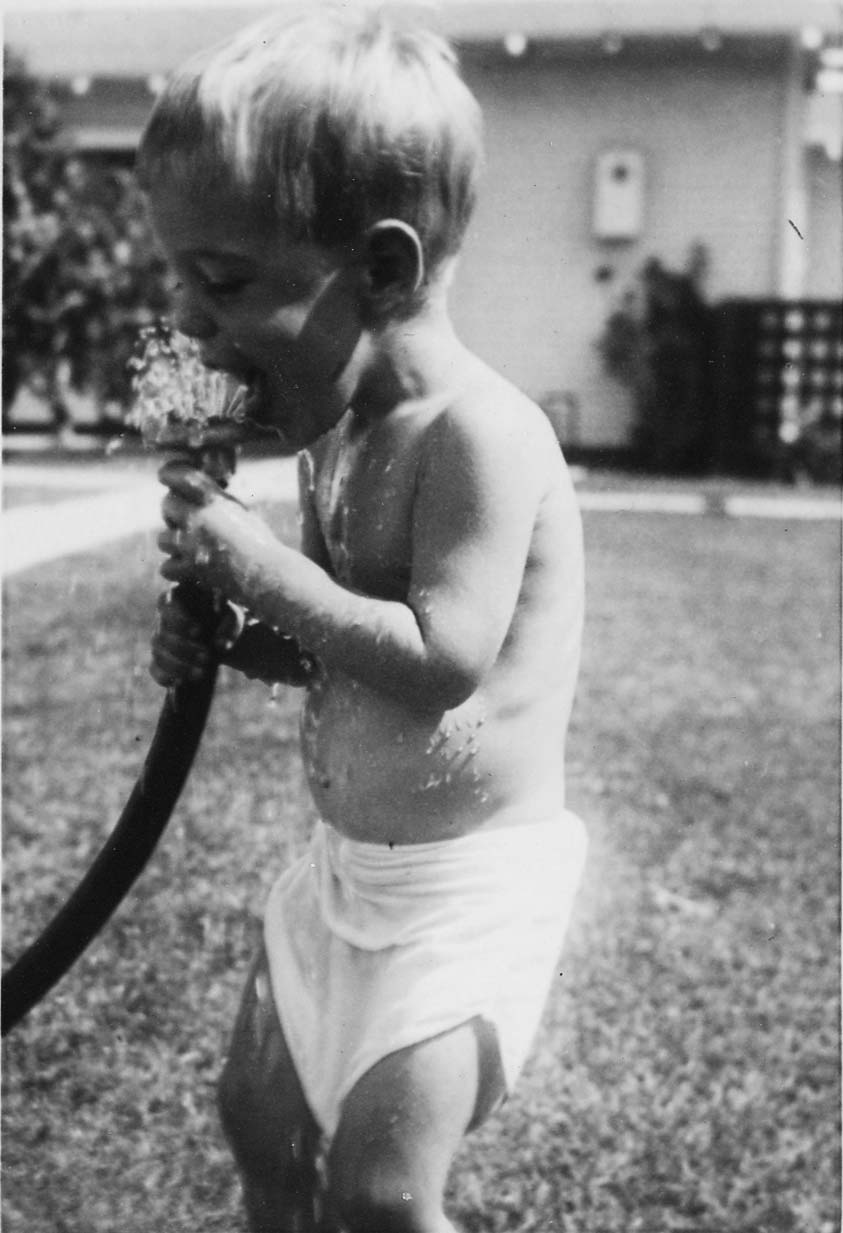 A candid photograph of a young toddler boy in a diaper playfully drinking water directly from a running garden hose in a grassy yard.