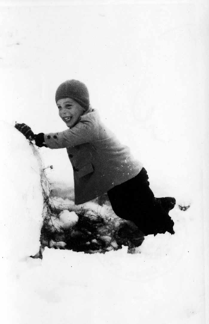 A young boy, Don Hall Jr., wearing a winter coat and hat, smiling as he pushes a giant snowball across a snowy field.
