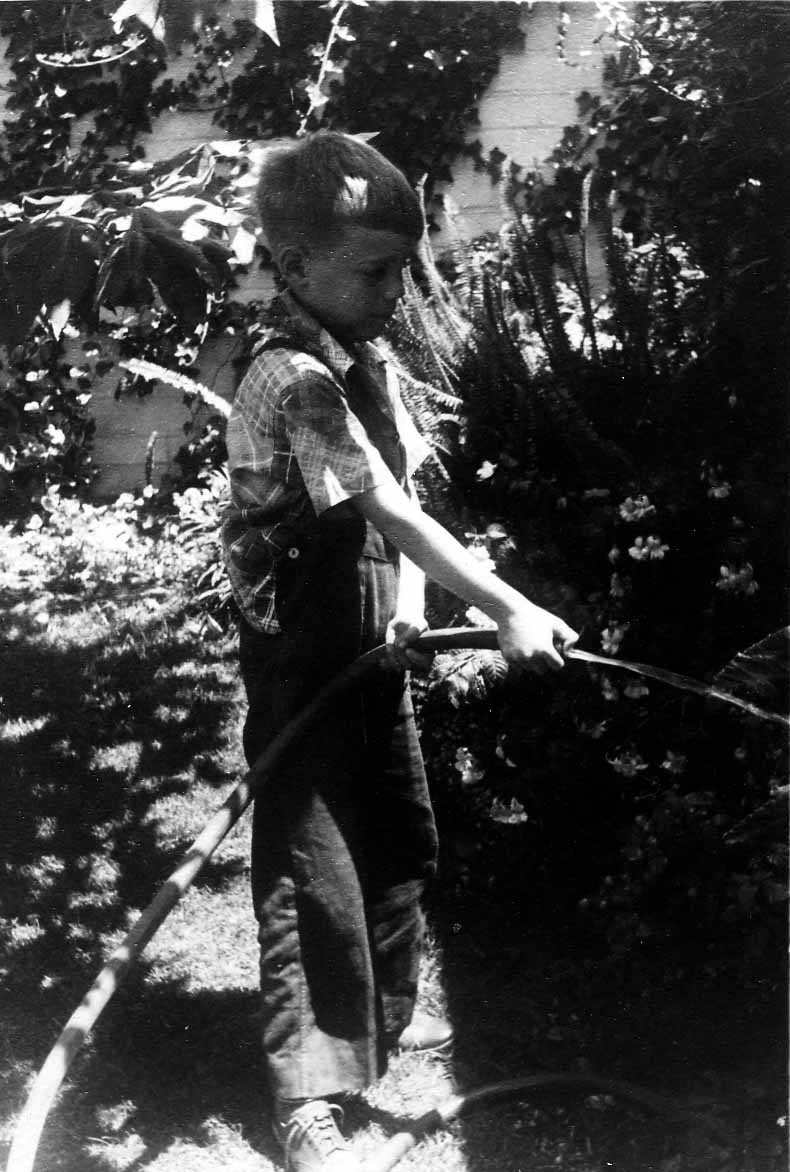 A young boy, Don Hall Jr., standing in the yard wearing overalls and a plaid shirt, holding a running garden hose to water the plants.