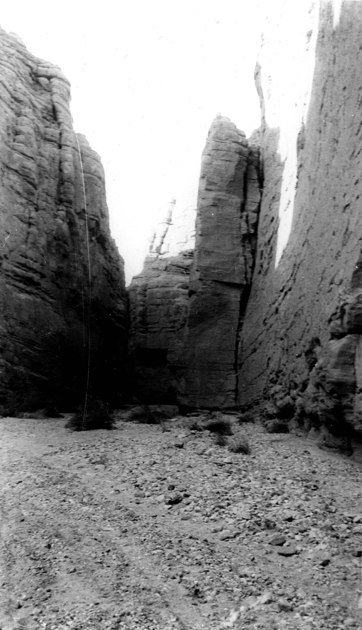 A tight view looking into a deep, narrow canyon passage flanked by sheer, weathered rock walls under bright overhead sunlight.