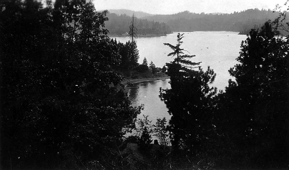 A serene view of a calm alpine lake framed by dark, framing foliage along the shoreline under an open sky.